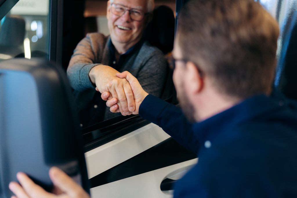 Personen som sitter i bilen är den äldre mannen med glasögon och grått hår, och han ler medan han sträcker ut handen för ett handslag. Personen som står utanför bilen är yngre, har kort brunt hår och bär en mörkblå skjorta. De skakar hand genom det öppna bilfönstret.