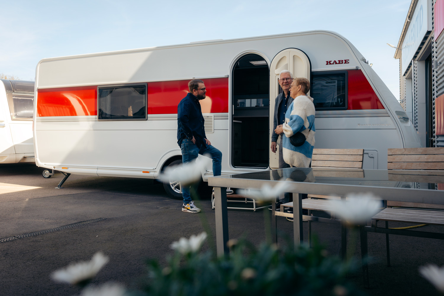 Tre personer står och pratar utanför en KABE husvagn vid Rudvi Fritid, en plats med skyltar synliga på byggnaderna. En äldre man och kvinna samt en yngre man i jeans och sneakers är vid husvagnens öppna dörr. I förgrunden syns ett bord och några blommor suddigt.