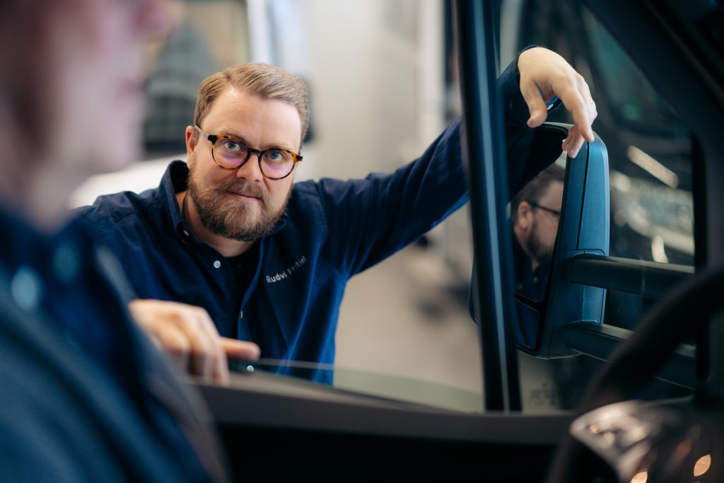 Viktor Rudvi lutar sig mot en bildörr och tittar mot en person i förgrunden. Han bär glasögon och en mörkblå skjorta med texten "Rudvi Fritid". Hans spegelbild syns i bilens sidospegel, och bakgrunden visar en ljus inomhusmiljö med fordon.