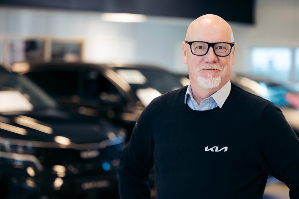 Micke Fransson, bilsäljare på Rudvi Bil, poserar i bilhallen iförd glasögon och en mörk tröja med Kia-logotyp.