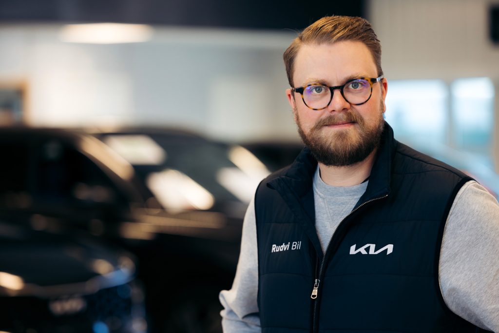 Viktor Rudvi, en man med glasögon och skägg, står inomhus i en bilhall med mörka bilar i bakgrunden. Han bär en mörkblå väst med texten "Rudvi Bil" och Kia-logotypen på bröstet.