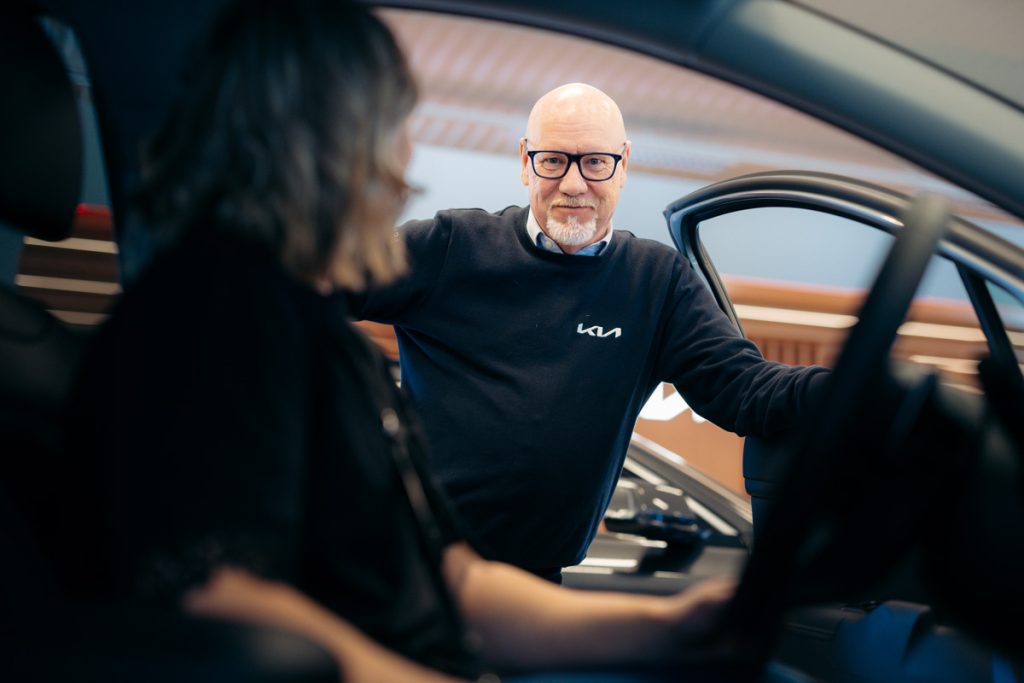 Micke Fransson från Rudvi Bil står vid en bildörr och pratar med en kund som sitter i förarsätet, inne i bilhallen.