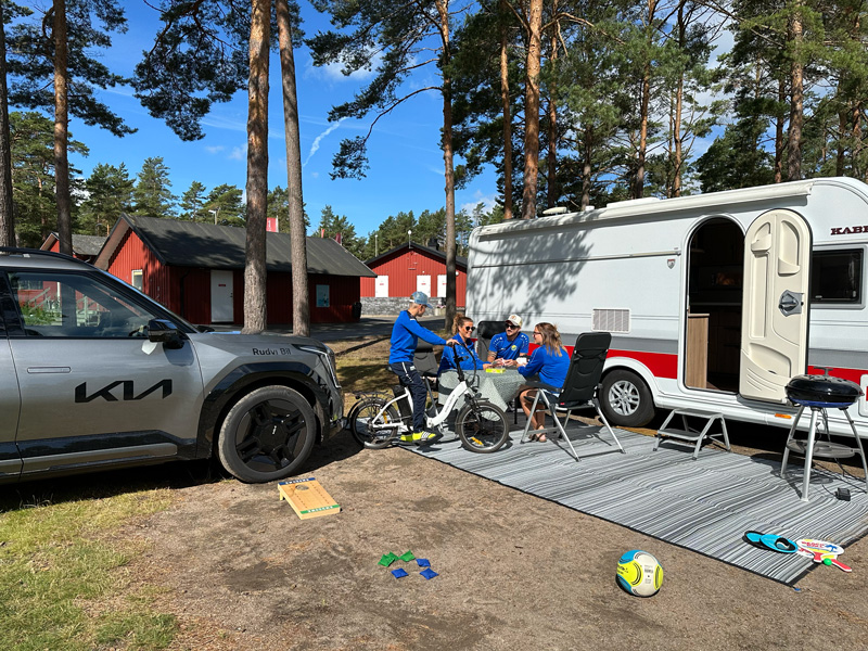 Familj campar med husvagn dragen av elbil – Kia EV9 parkerad vid campingplats med barn, elcyklar och fritidsaktiviteter i solen.