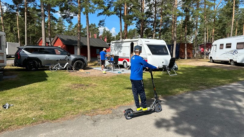 Camping med elbil och husvagn – barn åker elscooter, Kia EV9 parkerad bredvid husvagn på campingplats med elcyklar och familjeaktiviteter.